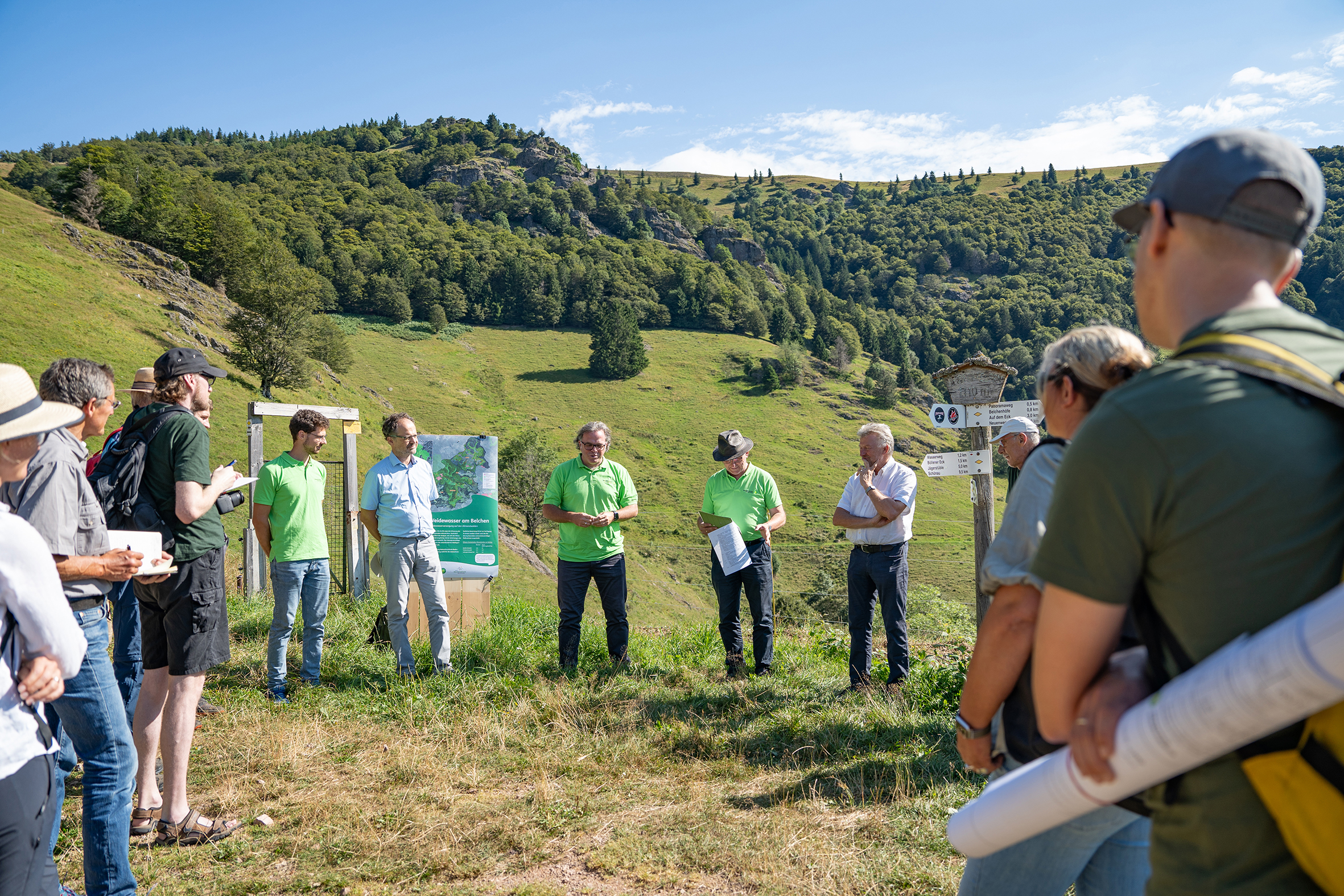 Eine Gruppe Menschen steht im Kreis vor einem Poster, im Hintergrund sieht man einen Berg, den Belchen